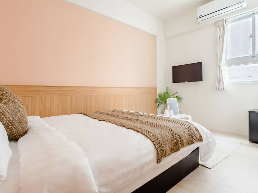 A hotel room with a bed, a flat-screen TV mounted on the wall, a window with curtains, and a small black refrigerator.