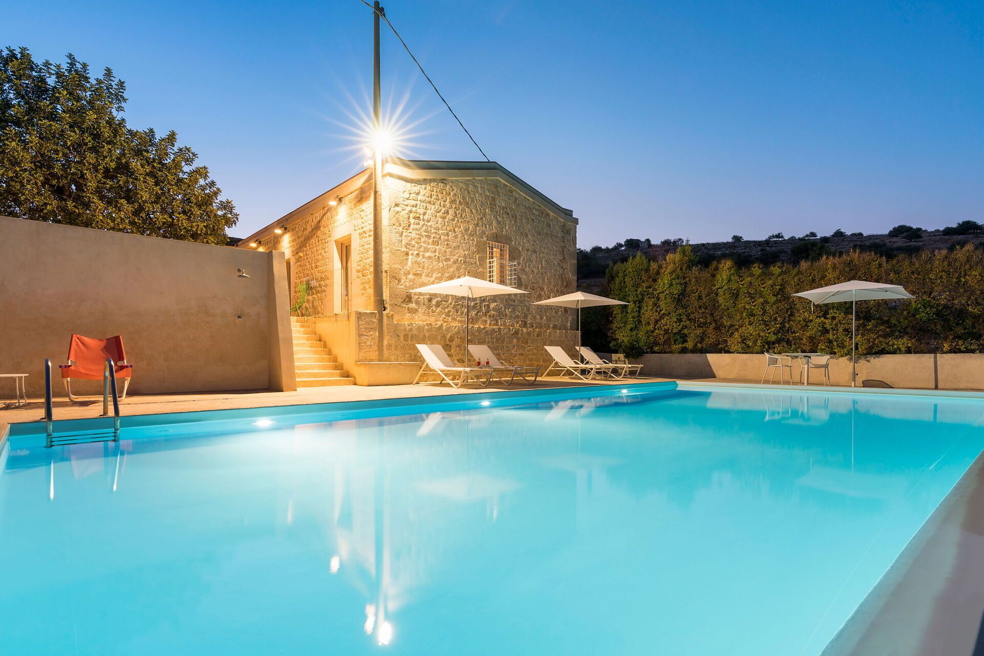 Uma piscina com espreguiçadeiras e guarda-sóis, um edifício de pedra com uma escadaria e um céu claro.