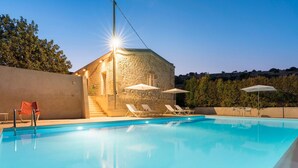 Uma piscina com espreguiçadeiras e guarda-sóis, um edifício de pedra com uma escadaria e um céu claro.