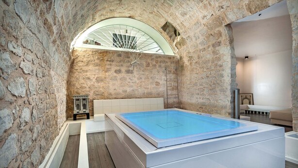 Um quarto de spa com parede de pedra, janela em arco de vidro, banheira branca e piso de madeira.