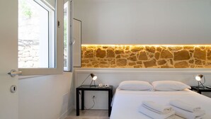 A bedroom with a stone wall, a bed with white linens, two bedside tables with lamps, and a window with a view of a stone wall.