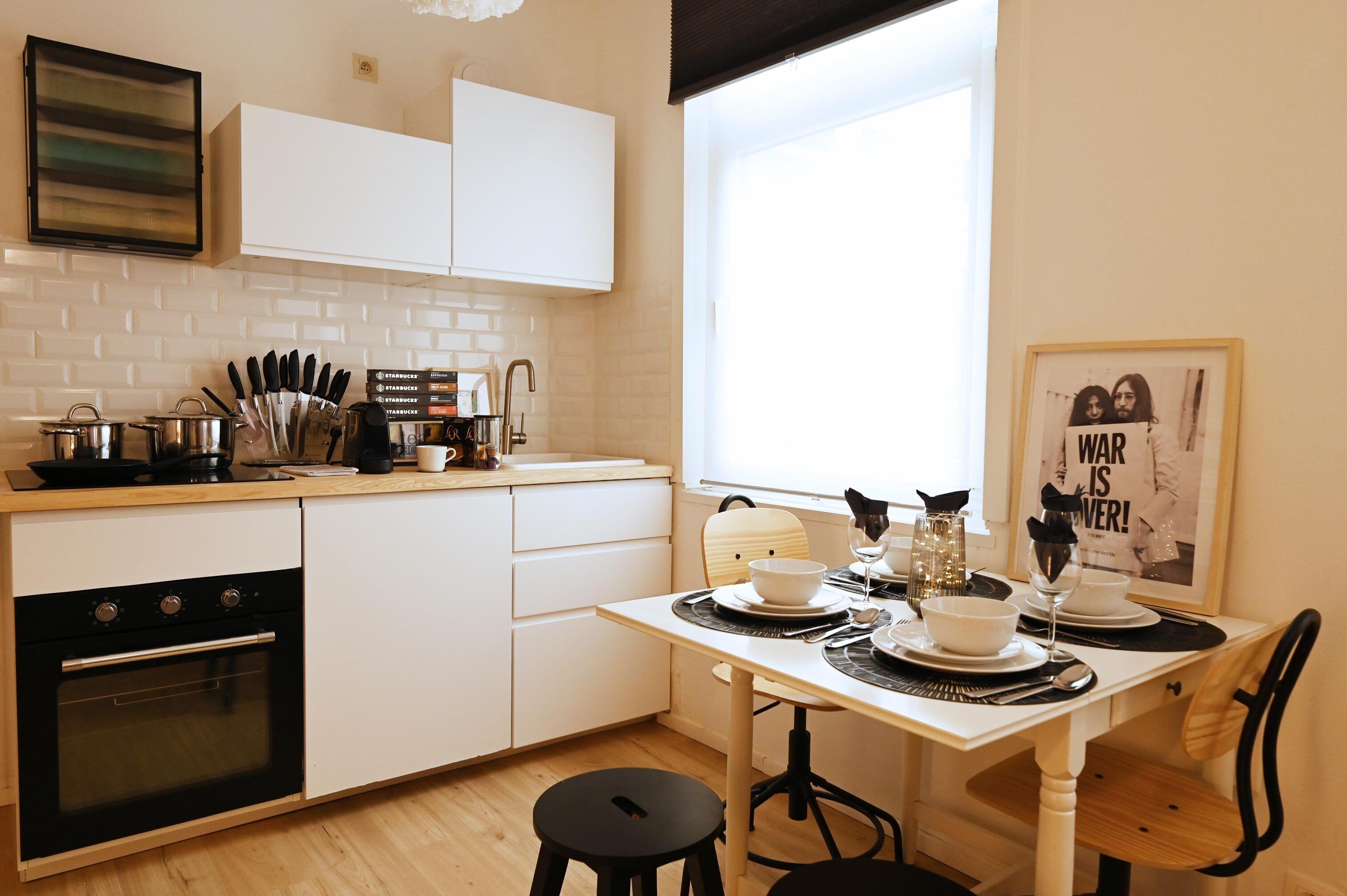Une cuisine moderne avec des meubles blancs, un four noir et une table à manger pour deux personnes, placée près d’une fenêtre offrant une belle vue.