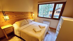 Een slaapkamer met een houten hoofdbord, een bed met witte lakens, een nachtkastje, een lamp, een raam met uitzicht op bomen en een radiator.