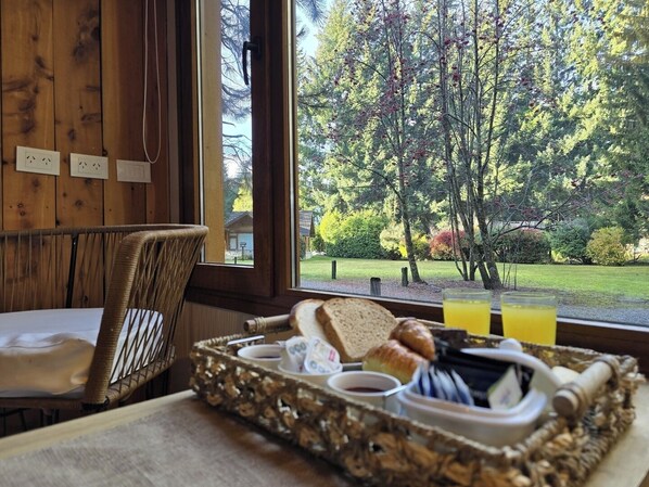 A breakfast tray with bread, butter, jam, and a drink on a table with a view of a forested area. - Remanso del Bosque - Maiten Cabin (Puerto Manzano)