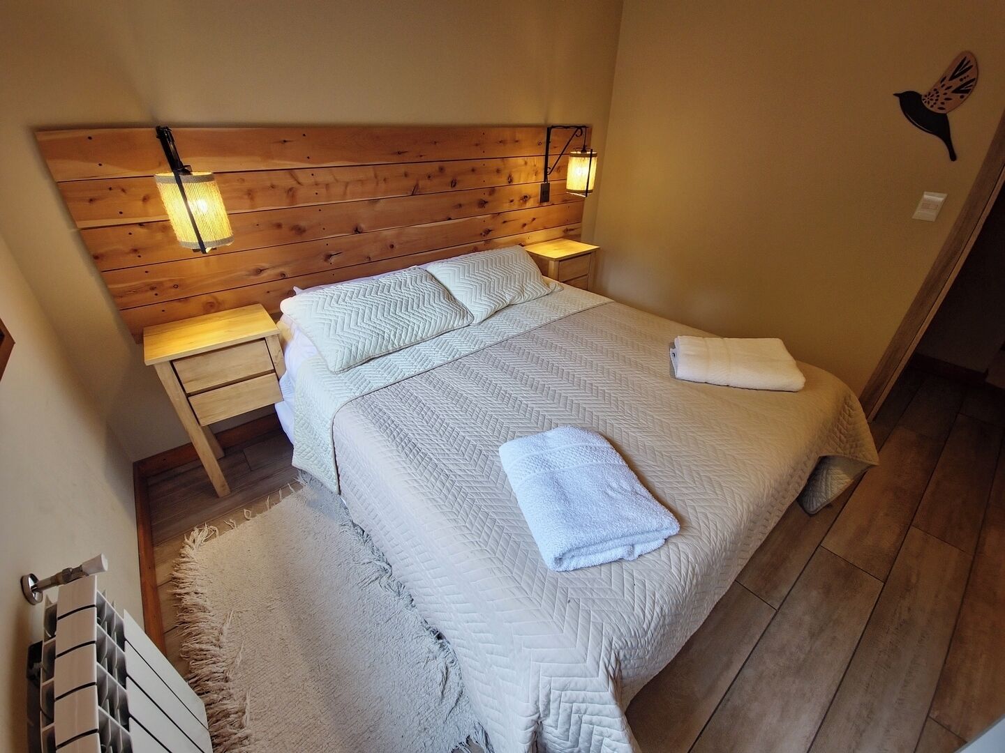 A bedroom with a wooden headboard, a bed with a quilted bedspread, two pillows, a nightstand with a lamp, and a wall-mounted bird decoration.