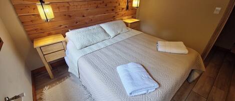 A bedroom with a wooden headboard, a bed with a quilted bedspread, two pillows, a nightstand with a lamp, and a wall-mounted bird decoration.