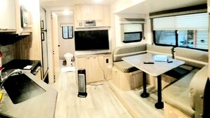 A compact RV interior with a kitchenette, a flat-screen TV, a small dining area, and a window providing a view outside. - The Magnolia House RV Winnebago Sleeps 3 (Lakeview)