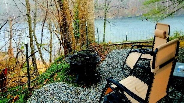 A secluded outdoor seating area with a view of a lake, surrounded by trees and a wire fence. - The Magnolia House RV Winnebago Sleeps 3 (Lakeview)