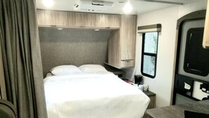 A compact RV bedroom with a bed, a small table, a window, and a kitchenette area. - The Magnolia House RV Winnebago Sleeps 3 (Lakeview)