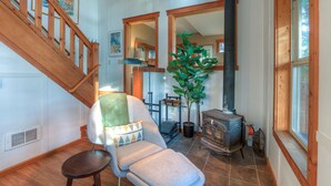 A cozy living room with a wooden staircase, a comfortable armchair, a large potted plant, and a wood-burning stove. - Cozy cabin in the woods, minutes from town, beaches, hiking (Eastsound)