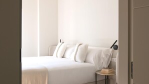 A minimalist bedroom with a bed, pillows, a bedside table with books, and a wall-mounted lamp. - HommyHome Duque de Veragua 101 (Sevilla)