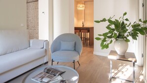A modern living room with a white sofa, a blue armchair, and a coffee table with a magazine. There is a potted plant on a side table and a dining area in the background. - HommyHome Duque de Veragua 101 (Sevilla)