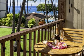 A wooden deck with a table set for two, featuring a bottle of champagne and glasses, surrounded by tropical flowers. - 🏖️Partial Ocean View Condo w/ A/C | Kona Isle B25 (Kailua Kona)