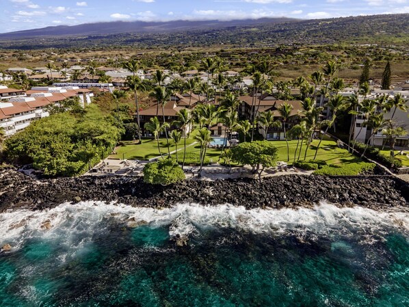 A coastal resort with clear turquoise waters, a rocky shoreline, and a view of a residential area with houses and palm trees. - 🏖️Partial Ocean View Condo w/ A/C | Kona Isle B25 (Kailua Kona)