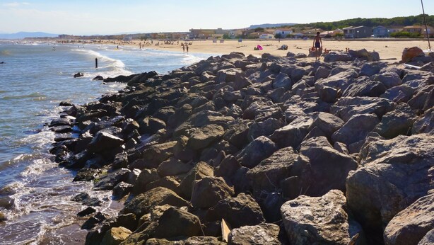 A rocky beach with waves crashing against the shore, a clear sky, and buildings in the background. - Maison des Pins Parasol by Interhome (Narbonne-Plage)