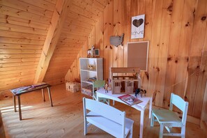 Ein gemütliches Dachzimmer mit Holzwänden, einer geneigten Decke, einem kleinen Tisch, einem weißen Stuhl, einem Bücherregal und einem Puppenhaus.