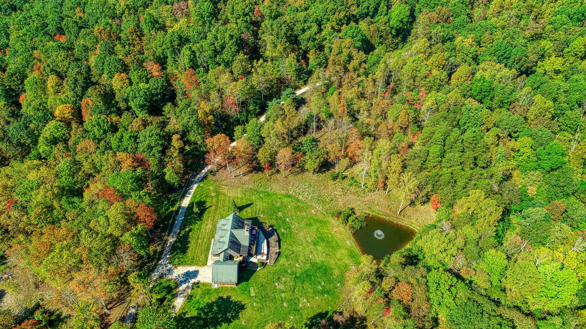 An aerial view of a property with a house surrounded by dense forest, a small pond, and a clear sky.