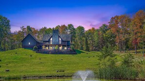 A large, rustic house with a prominent chimney, situated on a grassy hill with a pond in the foreground. - Wow! Timber Ranch Lodge, 43 acres, Sleeps 12, Family and Pet Friendly!  (Logan)