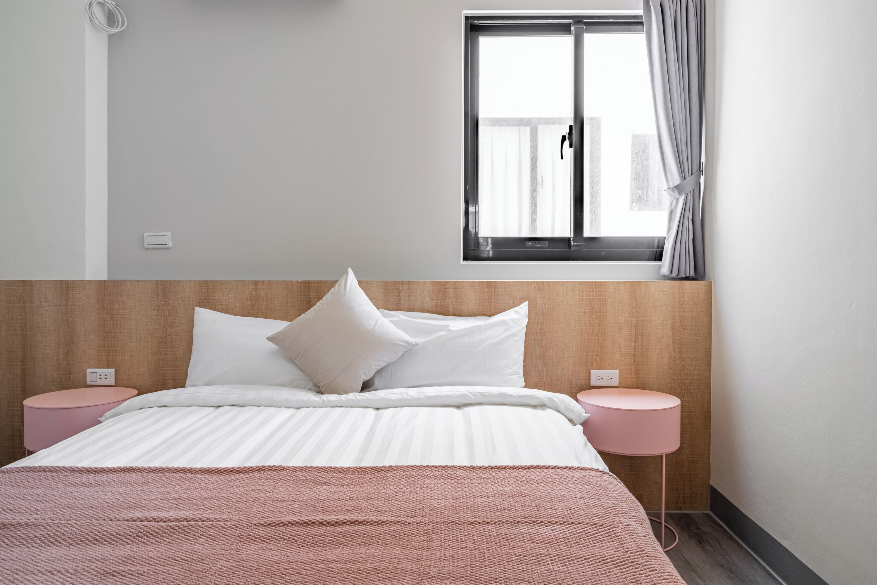 A neatly arranged bedroom with a wooden headboard, a large bed with white bedding, two pink bedside tables, and a window with curtains.