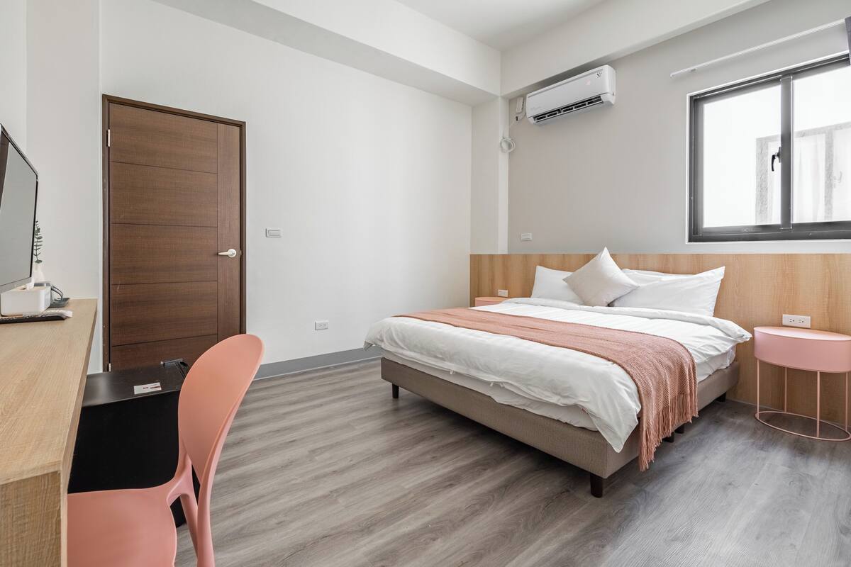 A bedroom with a bed, a desk, a chair, a bedside table, and a window.