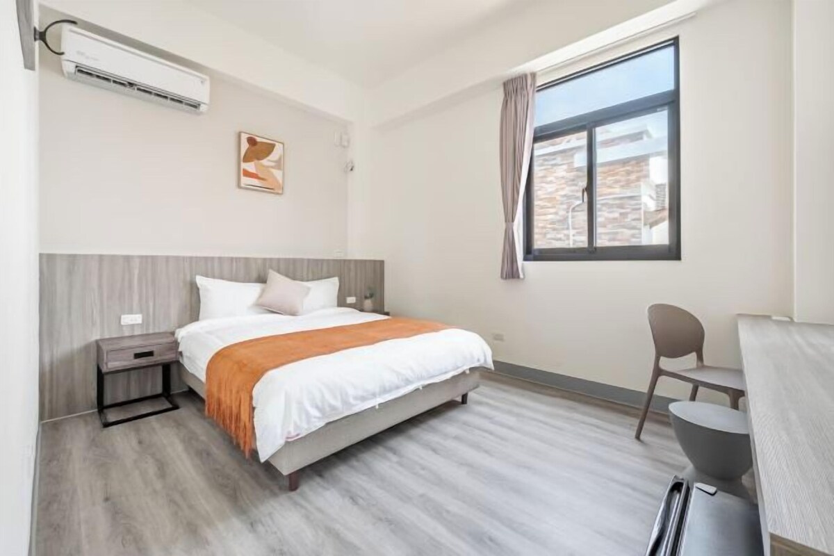 A modern hotel room with a bed, bedside table, and a desk with a chair.