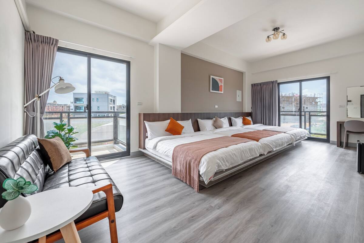 A modern hotel room with a large bed, a sofa, a dining table, and a view of the city through large windows.