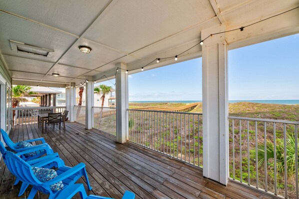 A covered outdoor area with wooden flooring, blue chairs, a table, and a railing overlooking a beach.