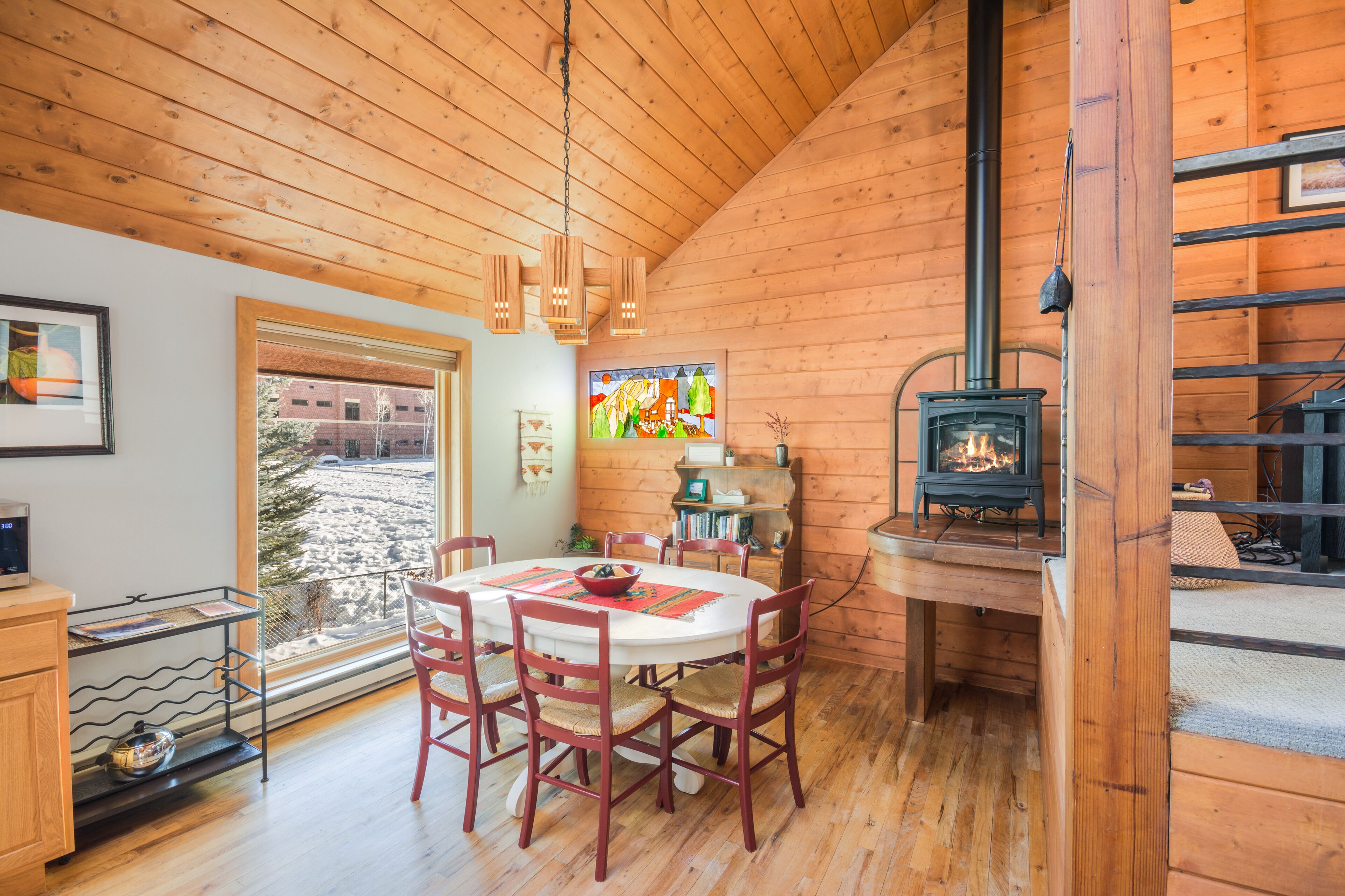 Interieur van een houten blokhut met een ronde eettafel, rode stoelen, een ingebouwde houtkachel en een groot raam met uitzicht op een besneeuwd landschap.