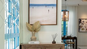 Un salon moderne avec un canapé blanc, une console en bois sur laquelle sont posés un vase et un livre, un tableau représentant une plage, le tout encadré, et un coin repas agrémenté d’un lustre.