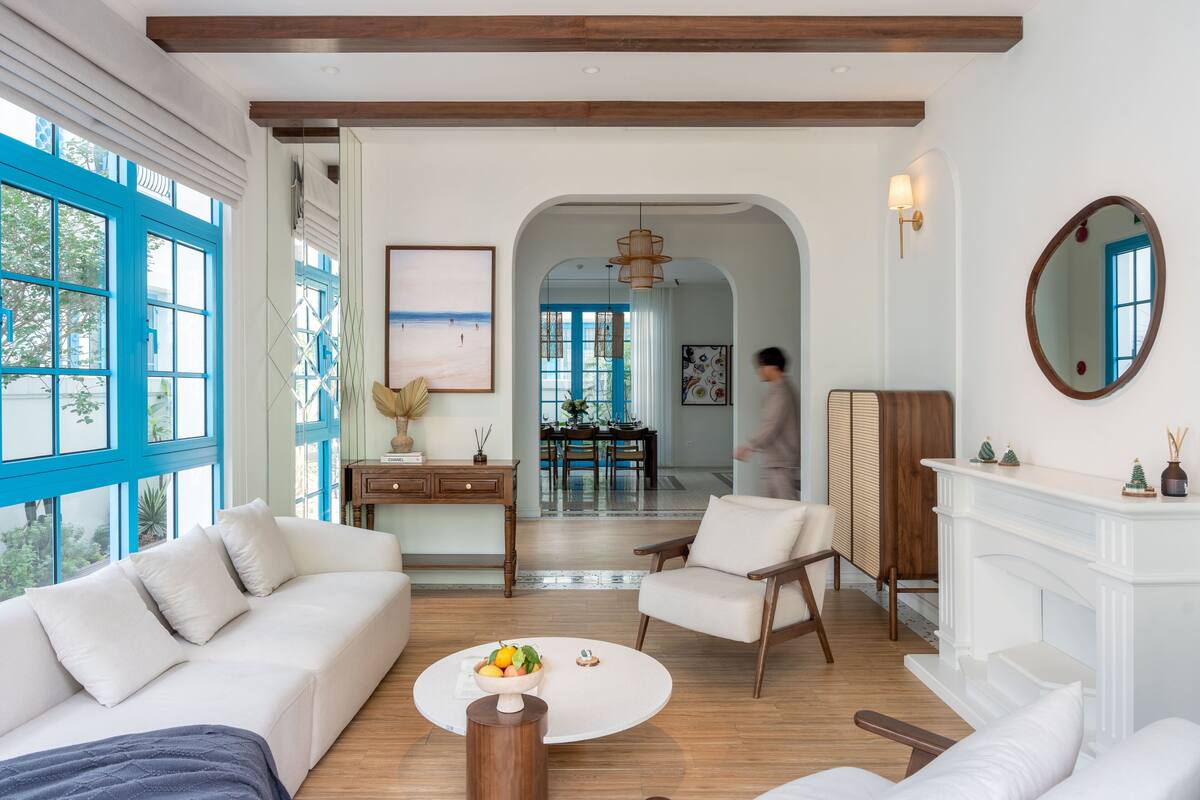 A spacious living room with a white sofa, a round coffee table with fruit, a fireplace, and large windows with blue frames.
