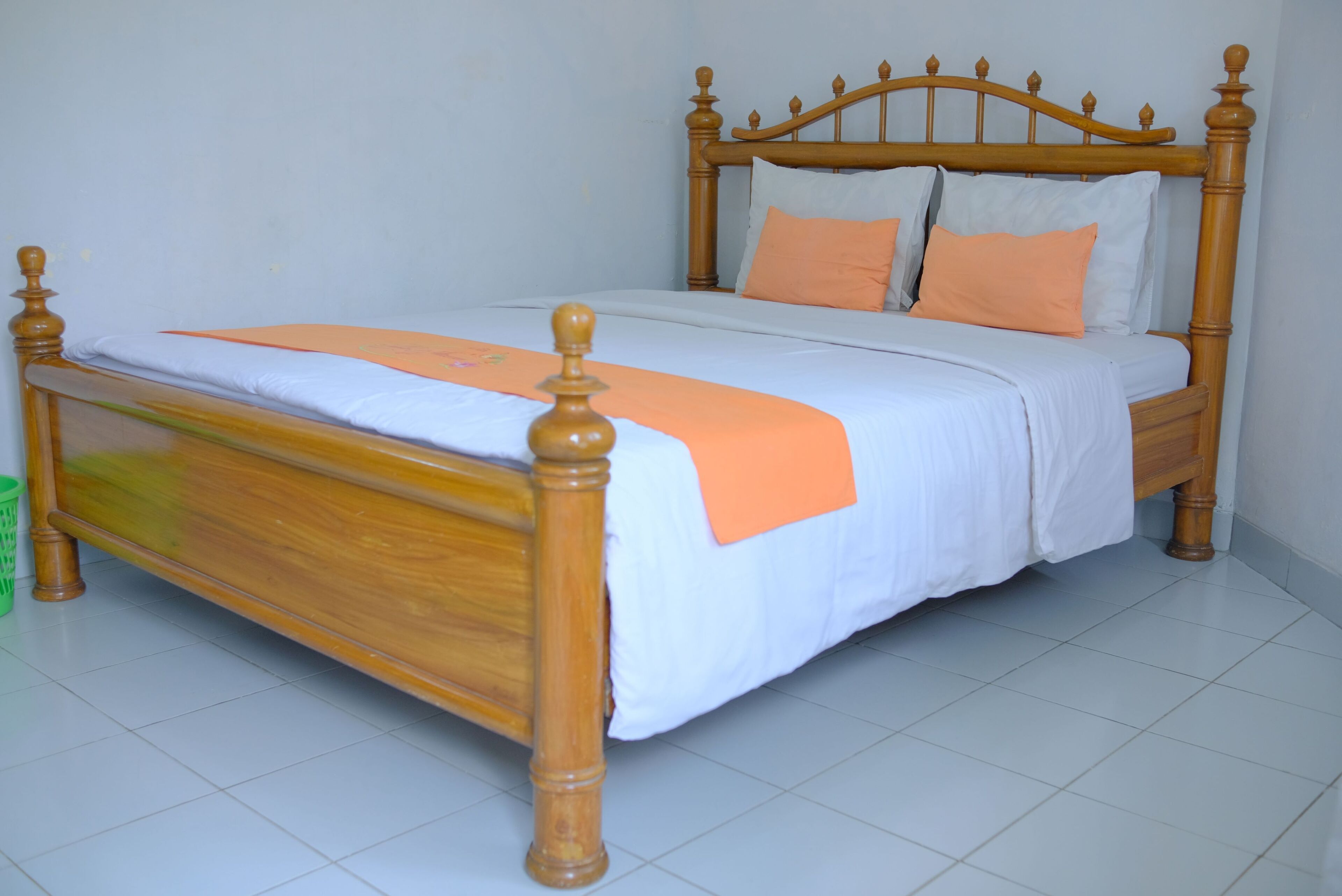 A wooden bed with a white and orange bedspread, a wooden headboard, and a green trash bin to the left.