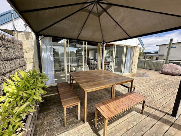 Un patio al aire libre con una mesa y bancos de madera, un paraguas grande y una puerta de vidrio que da a un espacio interior.