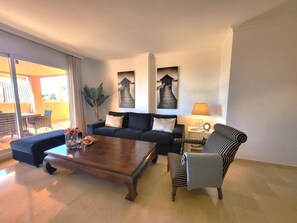 A living room with a sofa, armchair, and coffee table. There are two paintings on the wall, a lamp, and a plant.