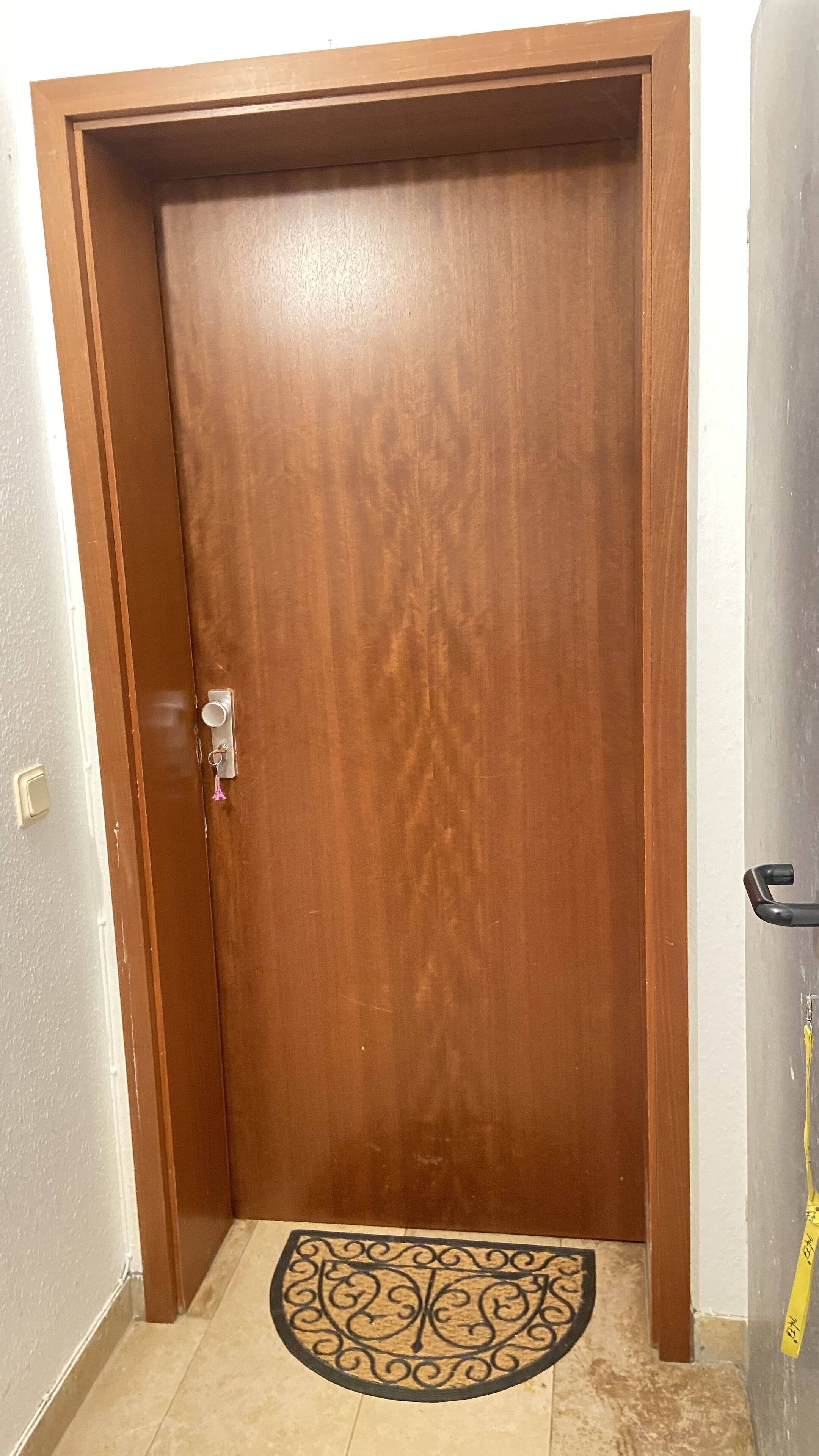Eine Holztür mit Türknauf und Schlüsselloch, eine Fußmatte mit dekorativem Muster und ein gelbes Klebeband an der Wand.