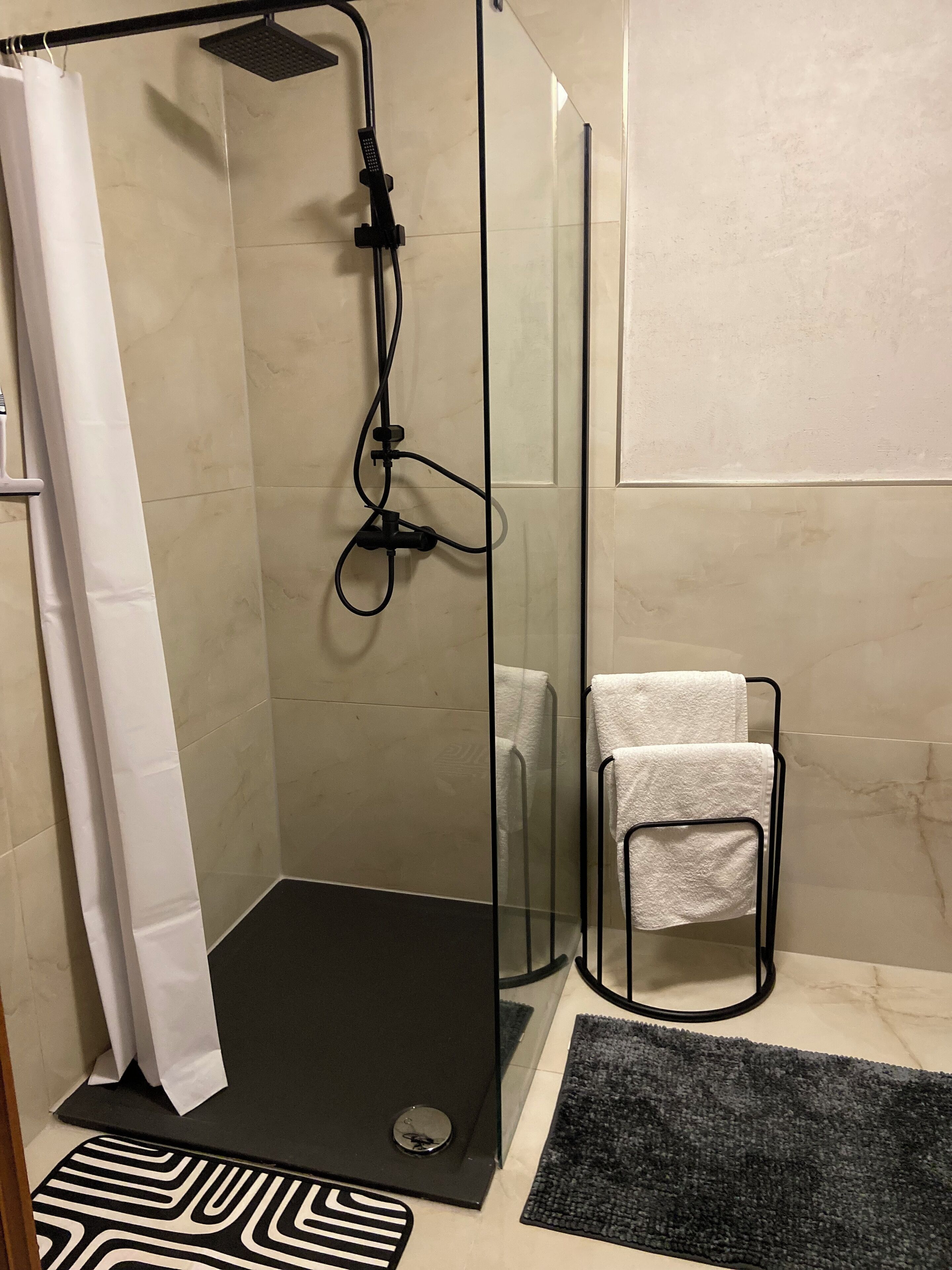 A glass-enclosed shower area with a black floor, a white towel rack, and a black and white patterned rug.