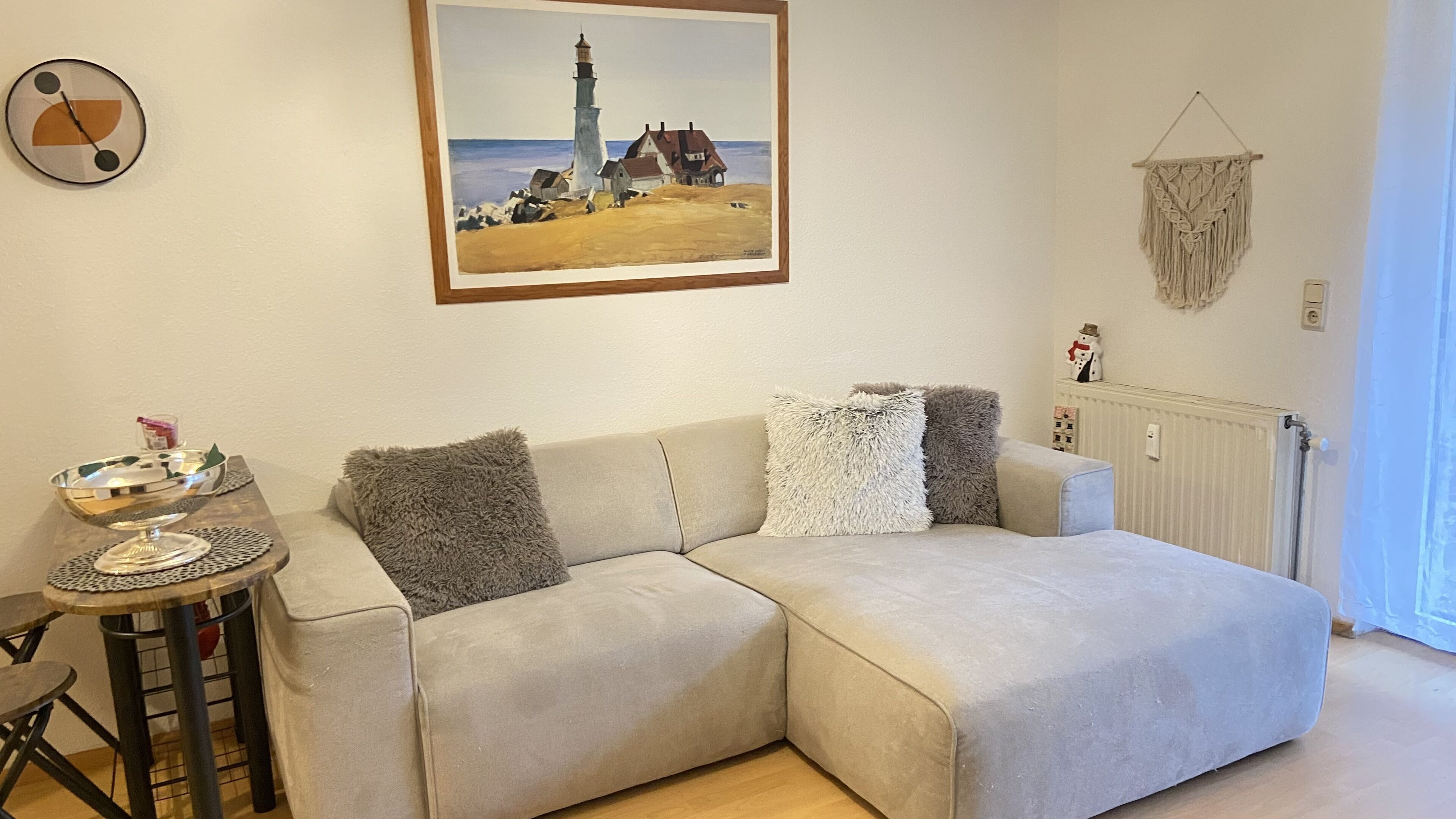 Een woonkamer met een beige bank, een lijstwerk met daarin een afbeelding van een vuurtoren, een macramé wandversiering en een klein tafeltje met een decoratieve kom en glas.