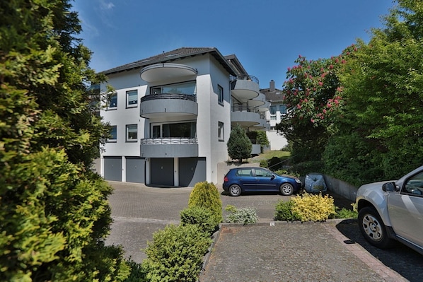 A modern multi-story residential building with balconies, a driveway, and parked cars.