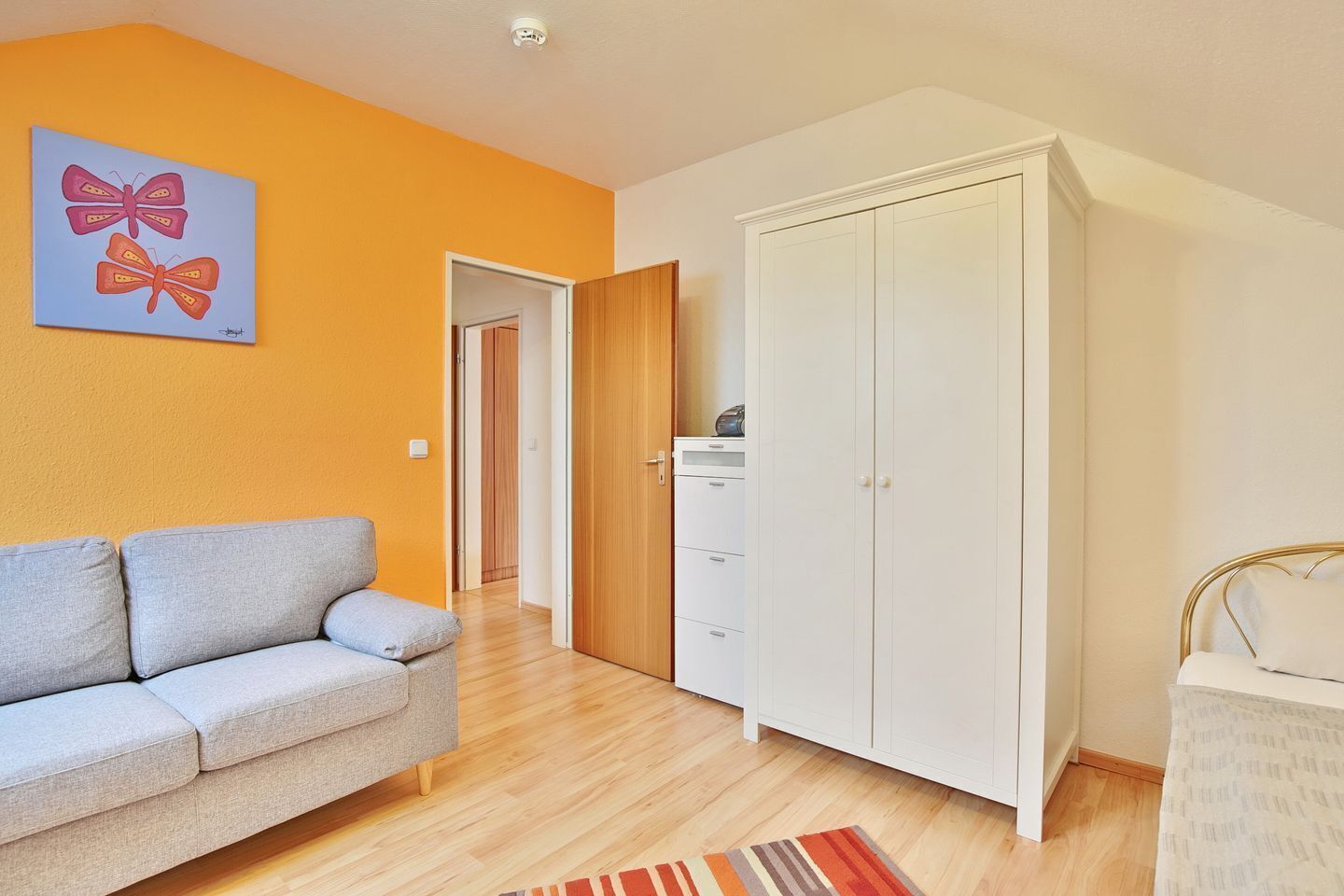 A room with a grey sofa, a white wardrobe, and a colorful rug.