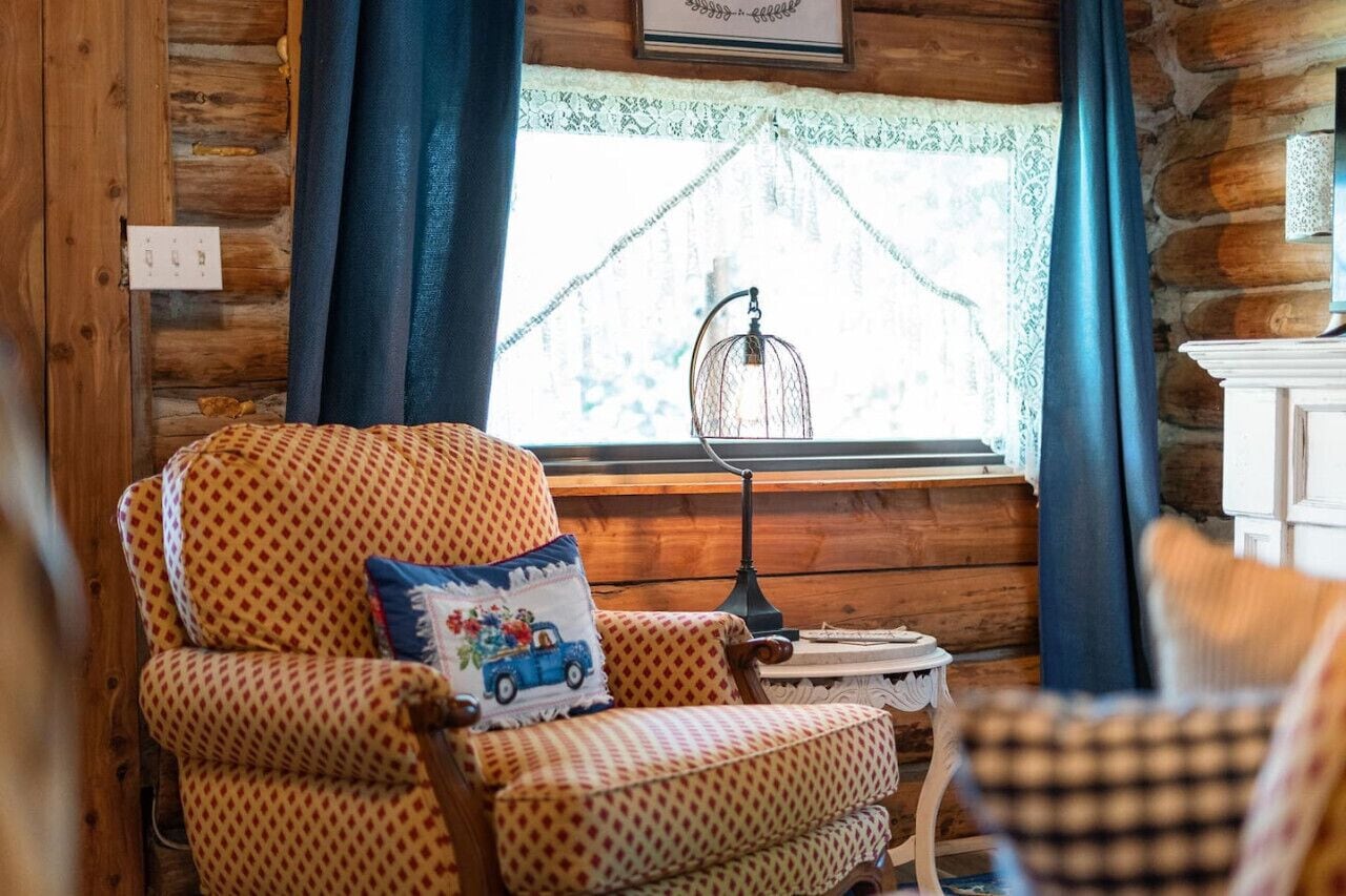 A cozy log cabin interior with a patterned armchair, a small table, and a window with a birdcage.