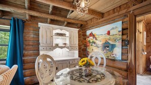 Ein rustikales Cabin-Interieur mit einer Holzwand aus Baumstämmen, einem weißen Schrank, einem Glastisch mit Stühlen und einem Gemälde einer ländlichen Szene.