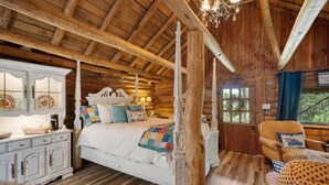 Ein Schlafzimmer in einer Blockhütte mit einem Himmelbett, einem weißen Frisierkommode und einem Fenster mit Blick auf grüne Natur.