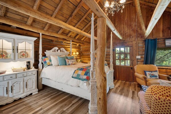 Une chambre dans une cabane en rondins, avec un lit à baldaquin, une commode blanche et une fenêtre donnant sur un paysage verdoyant.