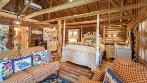 Ein rustikales Holzblockhaus im Inneren, mit Holzbalkendecke, einem Bett mit floralem Bettbezug, einer Couch mit gemusterten Kissen und einem Essbereich mit einem Schild, auf dem 'BISTRO' steht.