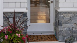 Una puerta blanca con un panel de vidrio, un cartel de bienvenida encima, un farol a la izquierda y una planta en maceta con flores rosas al frente.