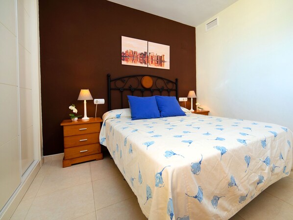 Une chambre à coucher avec un lit, deux tables de chevet, une lampe fixée au mur, une petite table avec un vase de fleurs et un tableau représentant un paysage urbain.