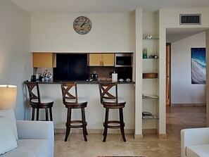 A modern kitchen with dark cabinets, a microwave, and a clock on the wall. There are three wooden stools and a white sofa in the living area. - Madeira Beach Yacht Club 247F by Distinctive Beach Rentals (Madeira Beach)