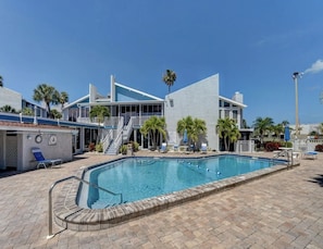 Uma área de piscina com piscina azul límpida, um espaço de estar coberto e um edifício de design moderno com palmeiras.