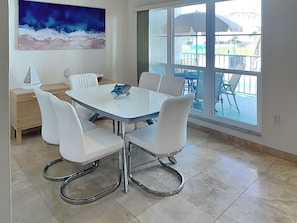Uma sala de jantar com uma mesa e cadeiras brancas, um quadro na parede e vista para o exterior através de grandes janelas.