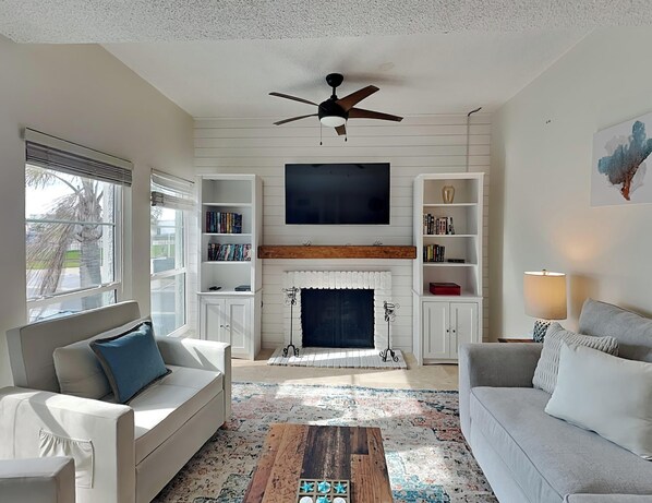 A living room with a fireplace, a ceiling fan, and a flat-screen TV mounted above it. - Madeira Beach Yacht Club 247F by Distinctive Beach Rentals (Madeira Beach)