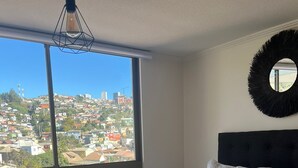 A bedroom with a large window, a bed with a patterned bedspread, a round mirror, and a hanging light fixture. - Hermoso Apartamento  en Viña del Mar (Viña del Mar)